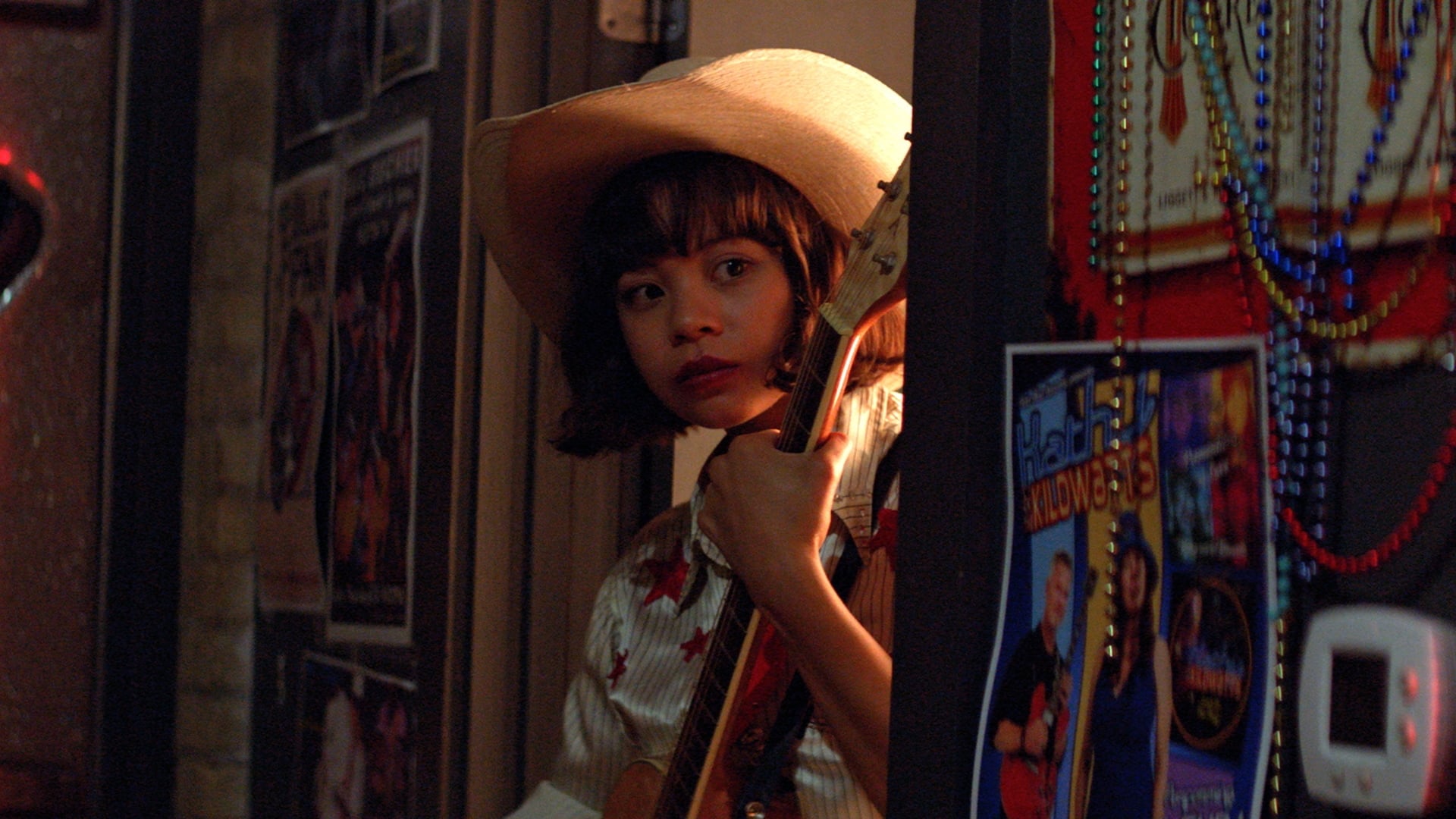 Image from the film 'Yellow Rose' (2019). A teenage girl peers out of a door. She wears a white cowboy hat and a striped button down shirt with red stars. There is an acoustic guitar braced to her chest. On the walls outside the door hang various posters and beaded necklaces.
