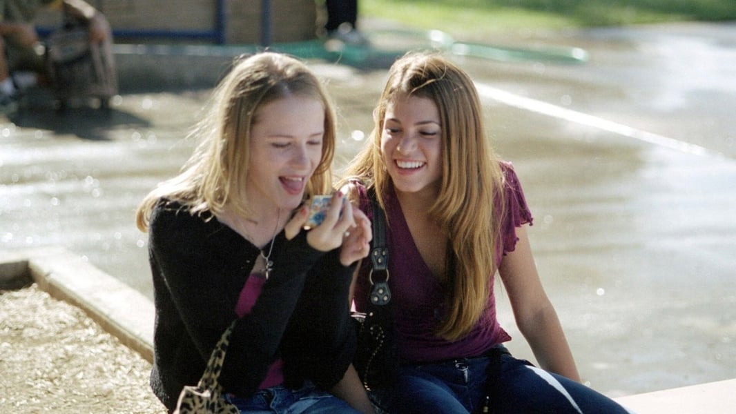 Evan Rachel Wood and Nikki Reed in a still from the film 'Thirteen'. The two girls sit outside wearing trendy outfits. Wood has her tongue out, and Reed is smiling as she looks over.